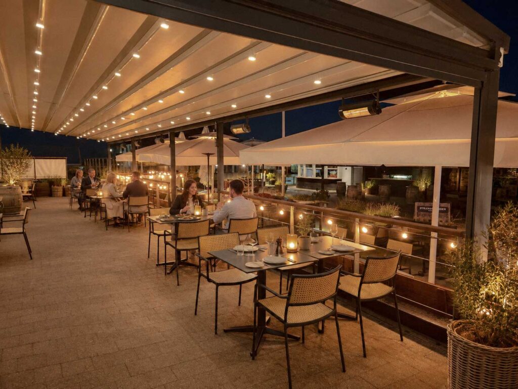 Restaurantens serveringsområde under et lyst pergolatag med glaspartier langs siden.