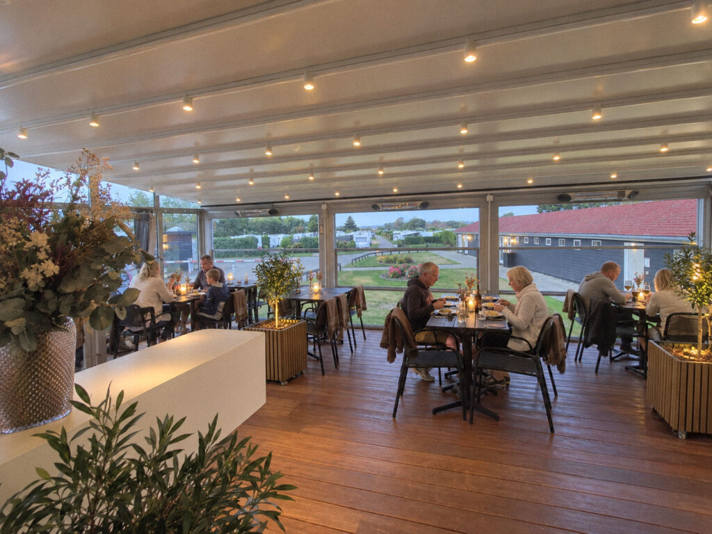 Fyldte restaurantborde under et langt, lyst pergolatag på en solrig dag.