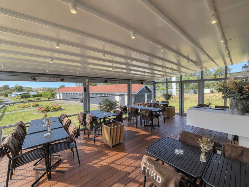 Caféborde og fletstole under et lyst pergolatag med spots og udsigt til grønne udearealer.