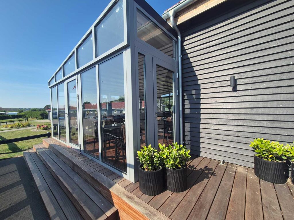 Mørk pergola med glaspartier ved en gavlbygning med vandret træbeklædning.