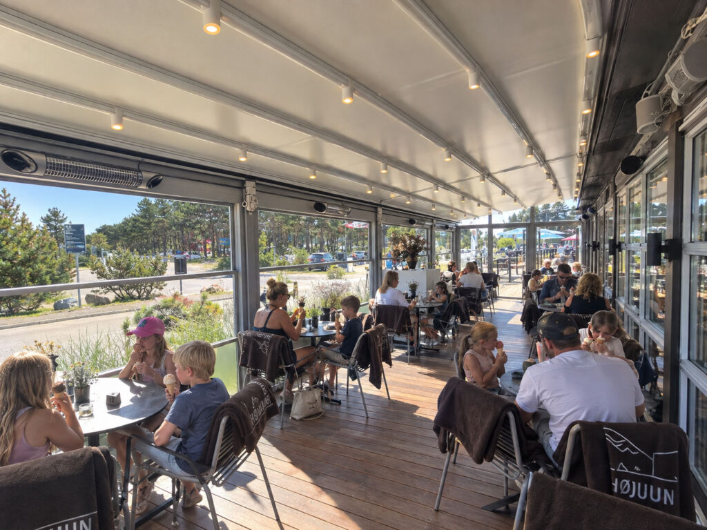 Gæster ved caféborde under en lang, lys terrasseoverdækning med planter langs kanten.