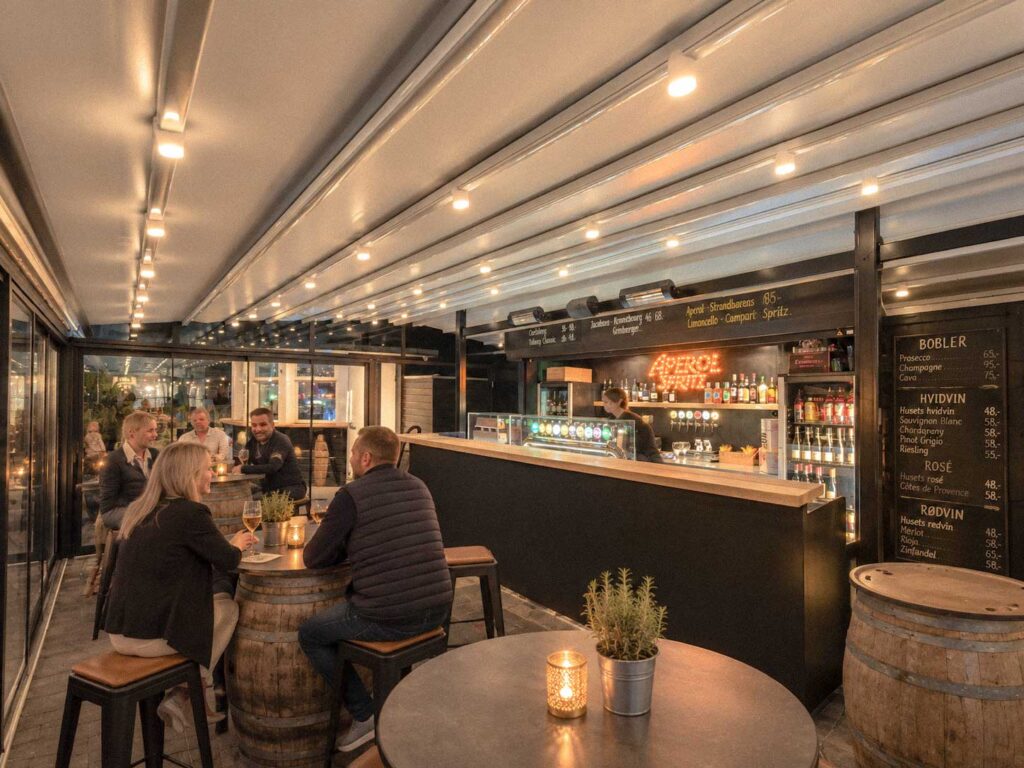 Mørk bardisk og serveringsområde under en lang, hvid terrasseoverdækning.