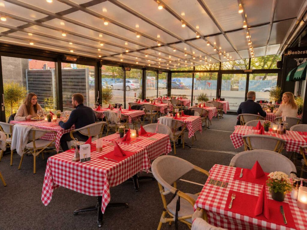 Gæster ved borde med rød-hvide duge under en lys terrasseoverdækning.
