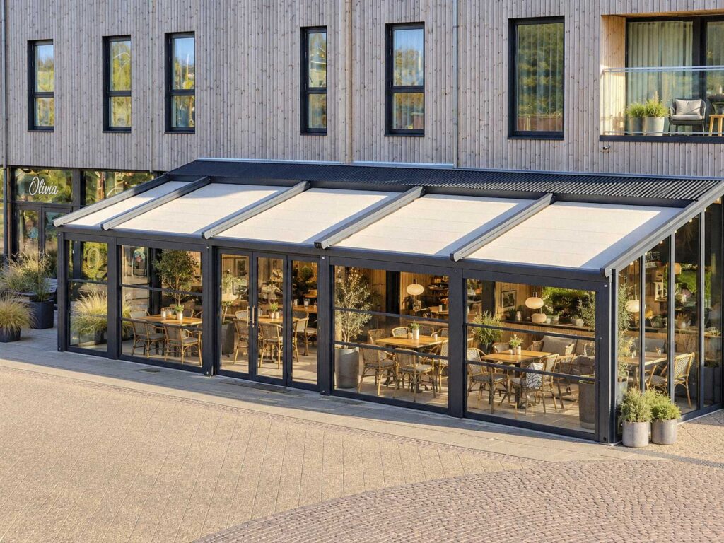 Glasafskærmet restaurantterrasse med pergola foran en hvid bygning i flere etager.