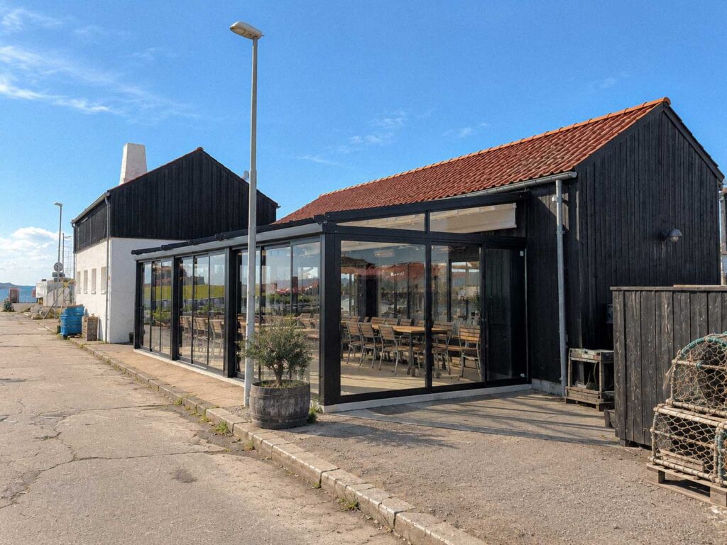 Mørk pergola med glaspartier ved en bygning med rødt tag og belægning foran.