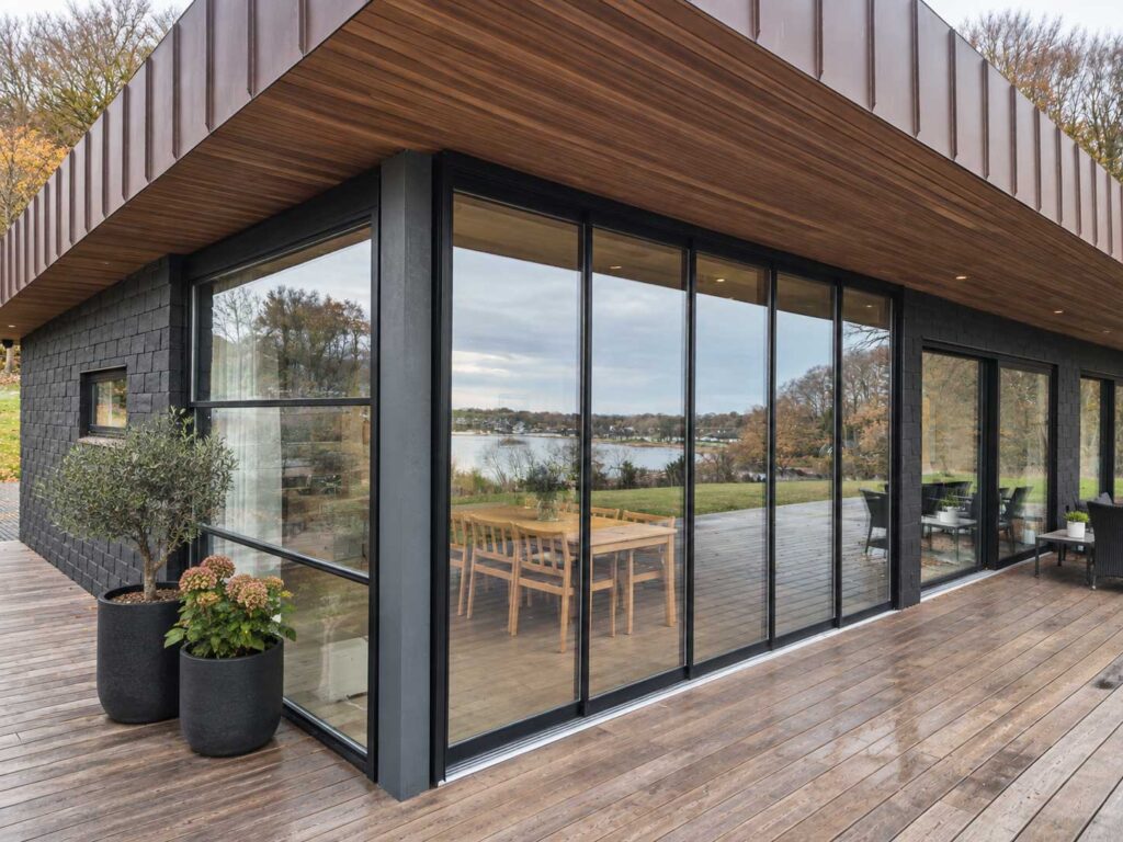Glasafskærmet terrasse med mørk pergola ved en bygning med vandret træbeklædning.