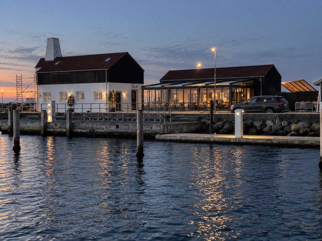 Restaurantterrasse med belyst pergola ved vandet, set fra den modsatte bred.