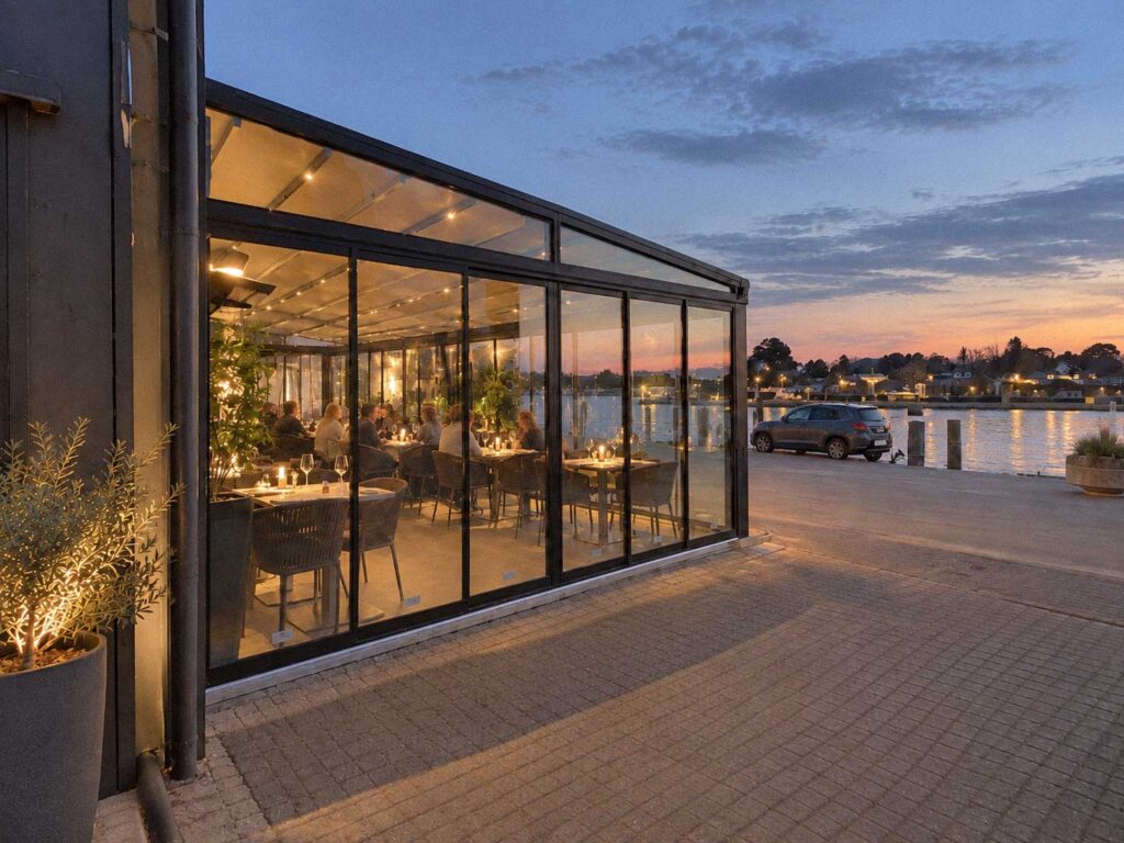 Mørk Ocean-pergola med glaspartier og belyst serveringsområde ved vandet om aftenen.