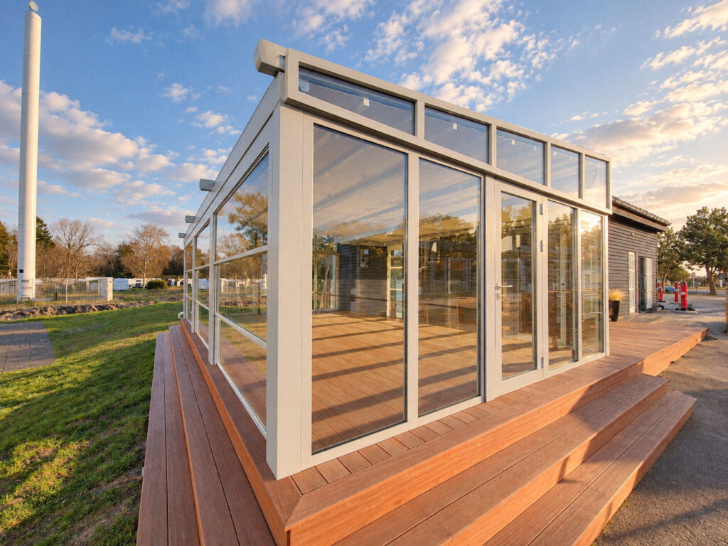 Hvid pergola med glaspartier omkring en terrasse med rødbrune terrassebrædder.