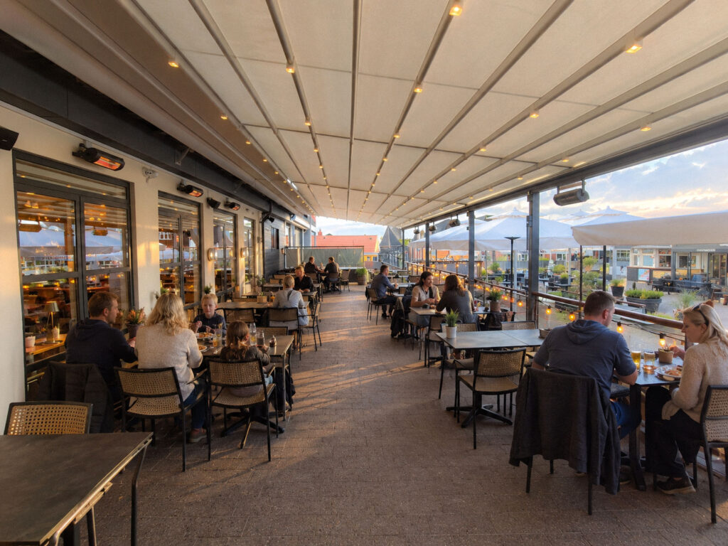 Restaurantborde under en lang, hvid terrasseoverdækning langs bygningens facade.