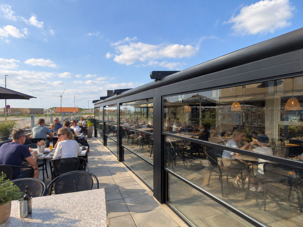 Mørk pergola ved Hvide Sande Røgeris hvide bygning, set fra hjørnet mod vejen.
