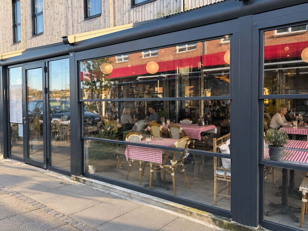 Mørk Ocean-pergola foran en café med mørk facade og rødt skilt mod gaden.