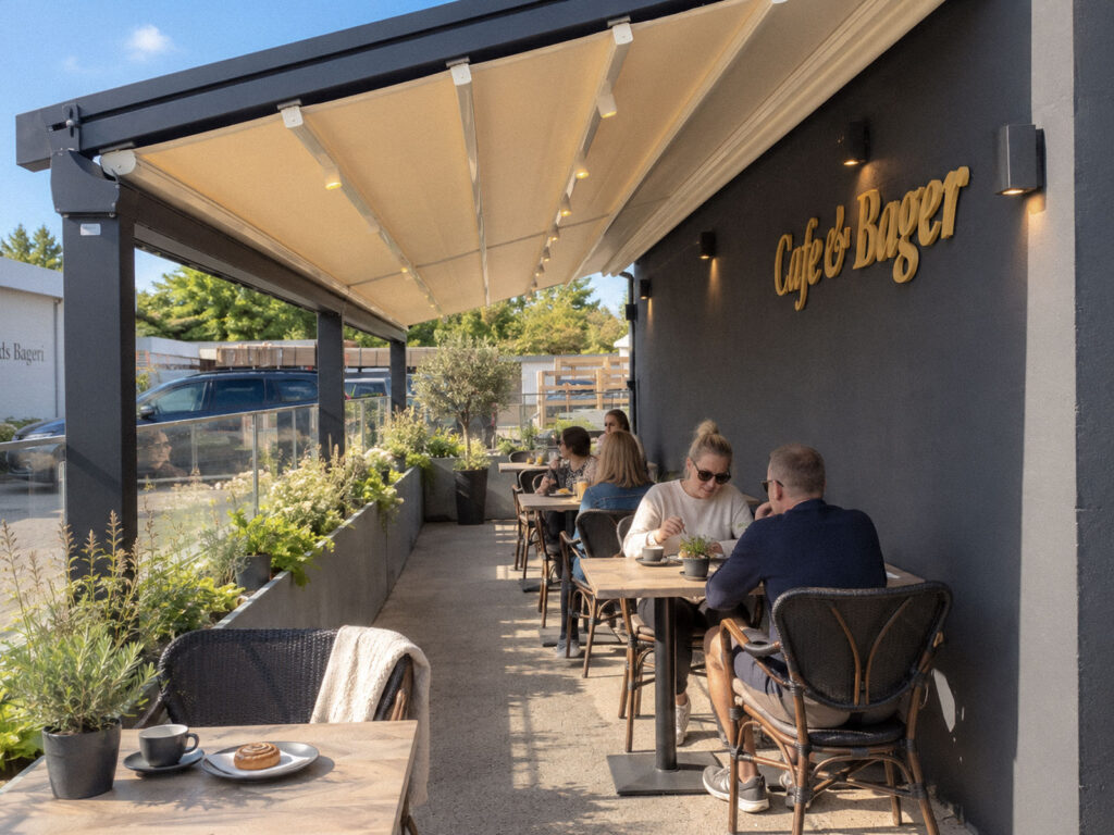 Gæster ved caféborde under et lyst Ocean-tag med belysning langs en mørk facade.