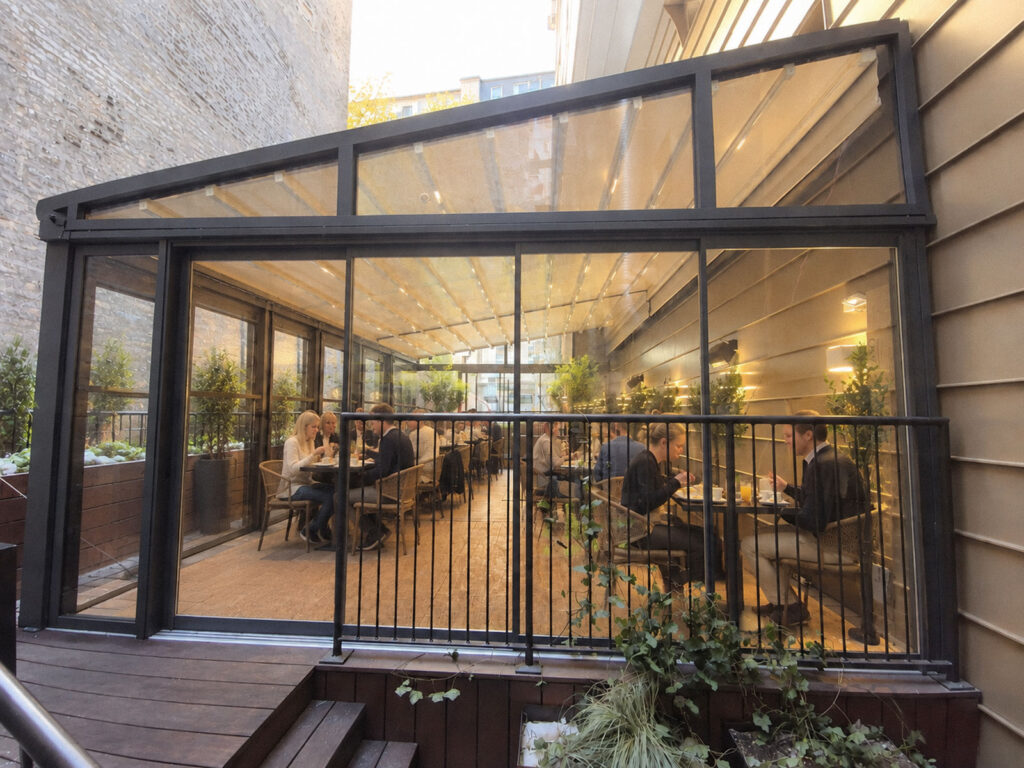Mørk Ocean-pergola med belyst restaurantterrasse bag glaspartier i skumringen.