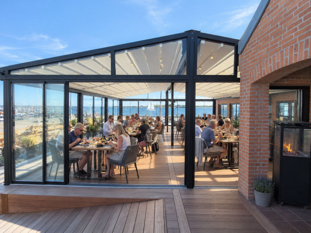 Mørk Ocean-pergola med glasfront og borde på en restaurantterrasse ved vandet.