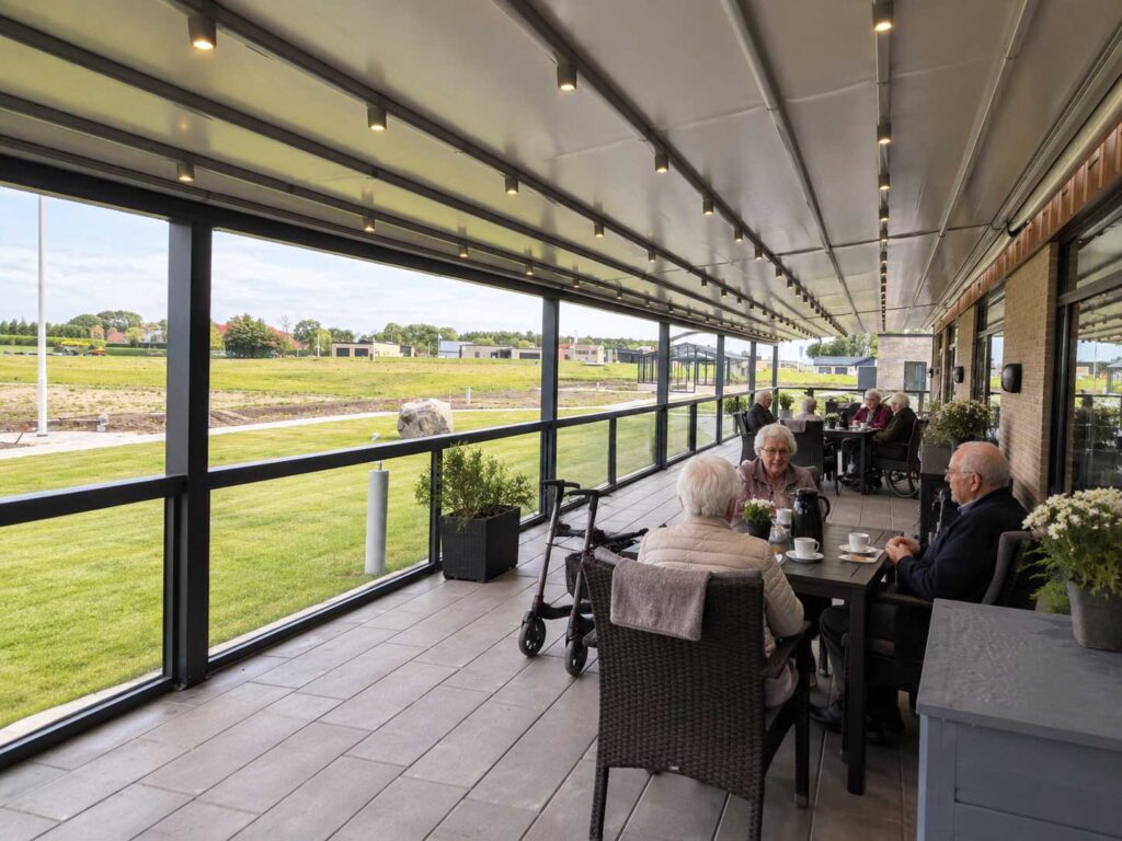 Borde og stole under en lang, lys terrasseoverdækning ved et plejehjem med udsigt til plænen.