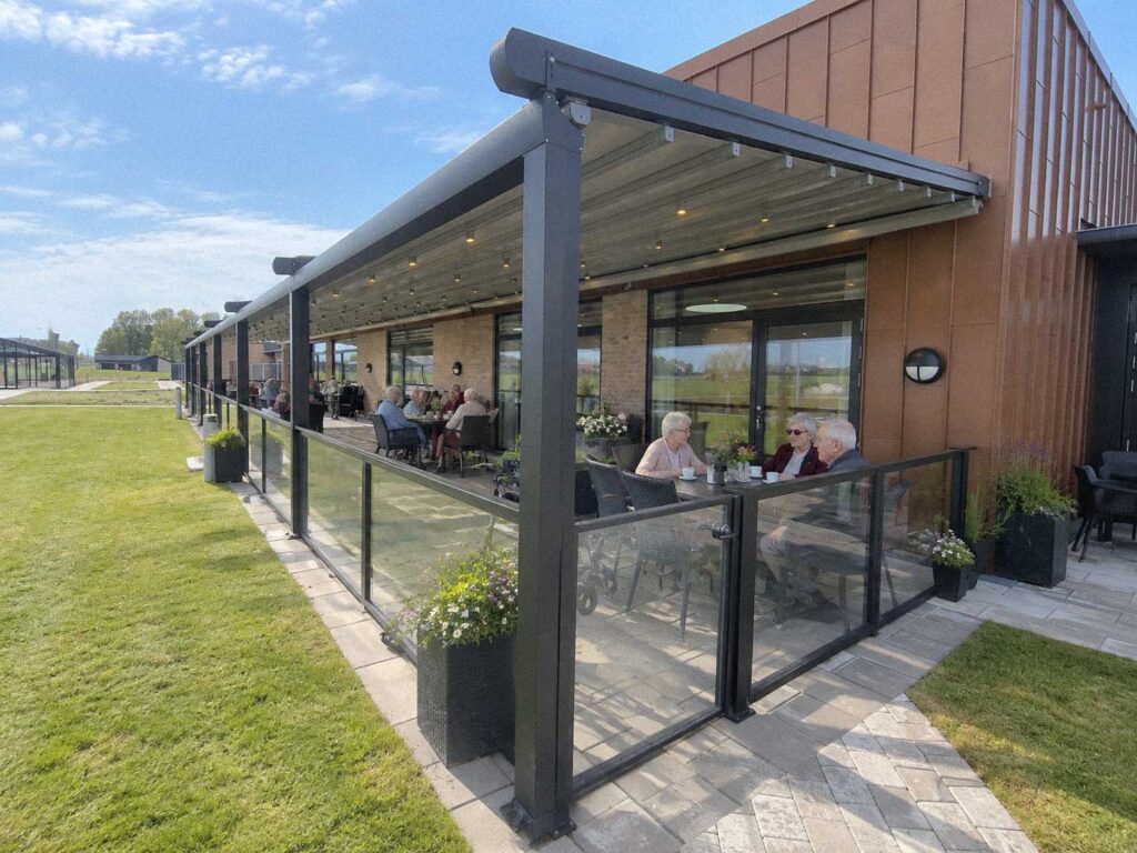 Mørk pergola ved et plejehjem med orange mursten, træbeklædning og græsplæne.