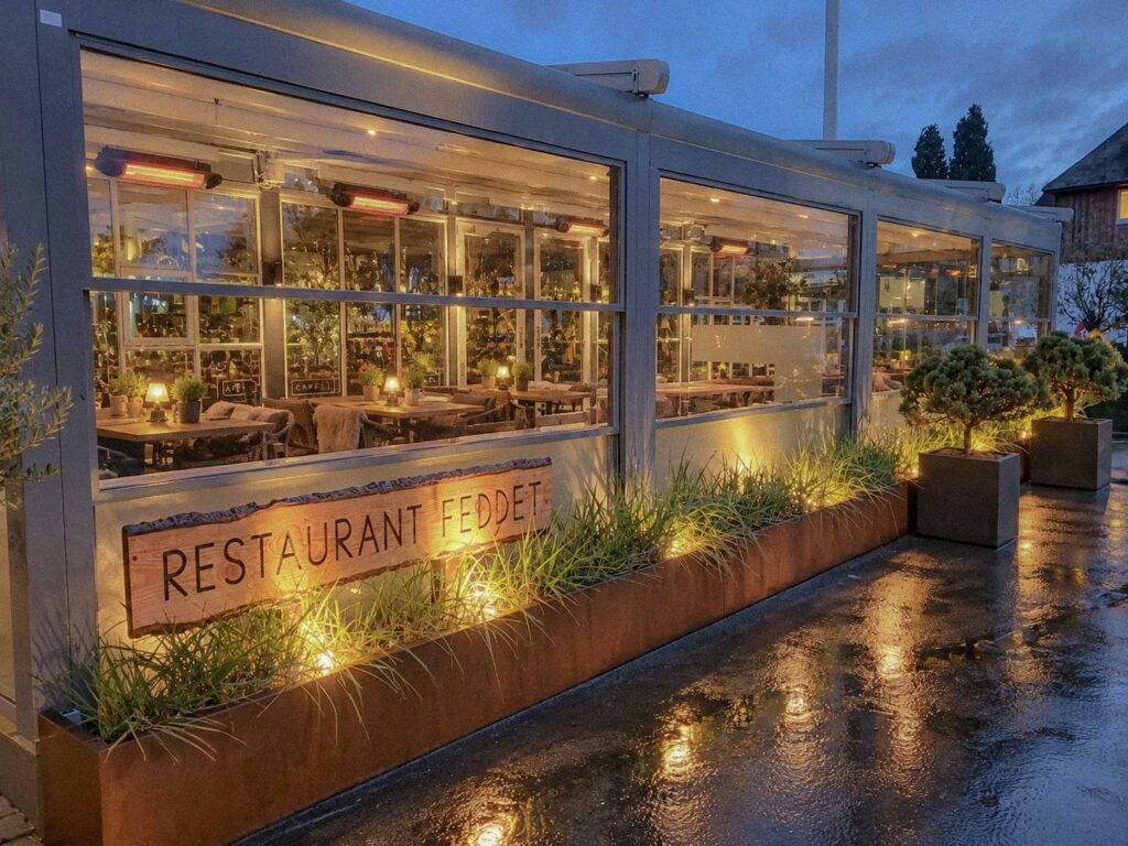 Restaurant Feddets glasafskærmede terrasse med mørk pergola og tændt belysning om aftenen.