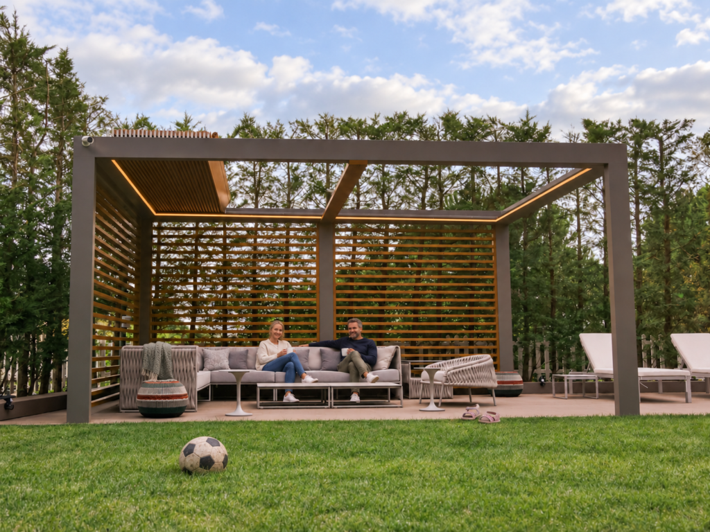 Pergola med åbent lameltag, trælameller og to personer i havens lounge